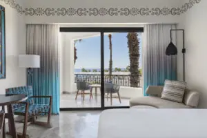 Habitación de hotel luminosa con muebles modernos, puertas corredizas que se abren a un balcón con sillas y vista al mar.