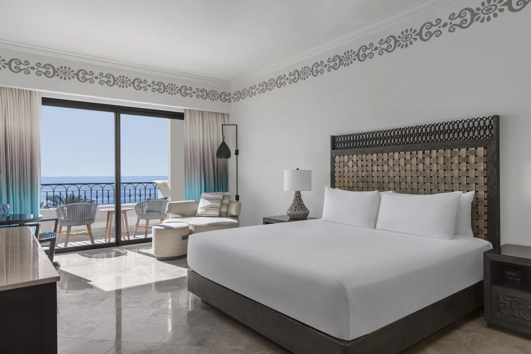 Modern hotel room with king bed, seating area, ocean view balcony, and elegant patterned décor.