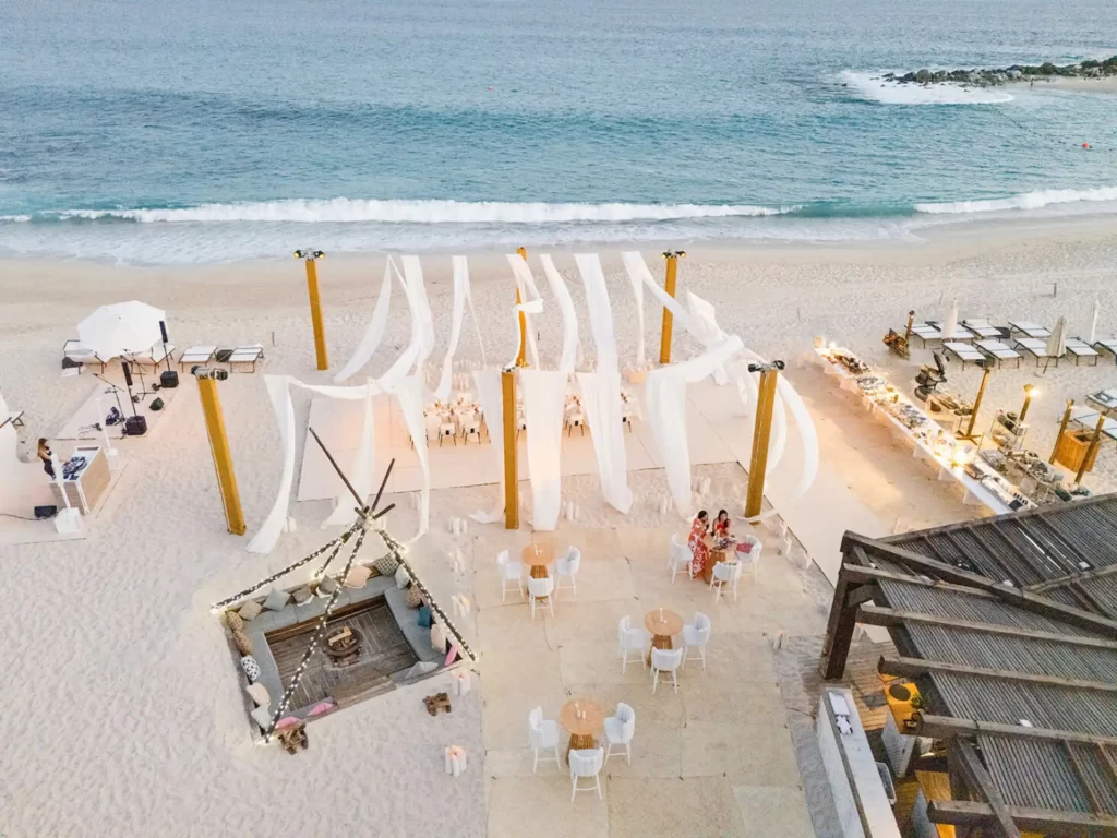 Elegant beachside wedding setup with flowing white drapery, tables, lounge area, and ocean backdrop.