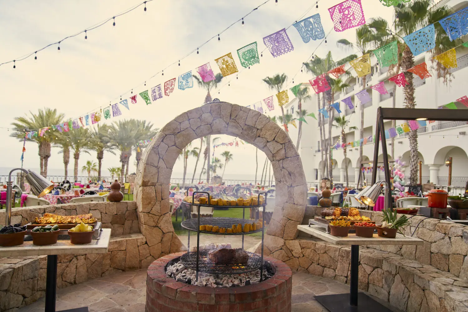 Outdoor Mexican fiesta setup with grill, colorful papel picado banners, palm trees, and festive food.