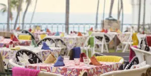 Colorful outdoor dining tables with pattern cloths and napkins, surrounded by decorated chairs, near ocean.