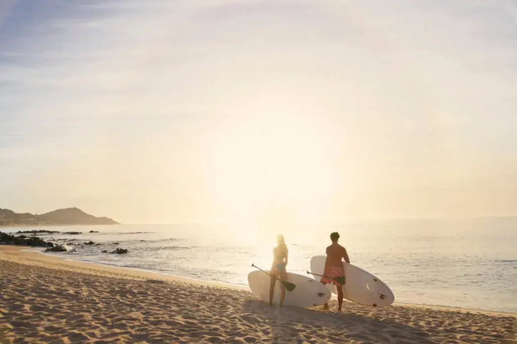 Two surfers carrying boards walk along a sandy beach toward the ocean at sunrise.