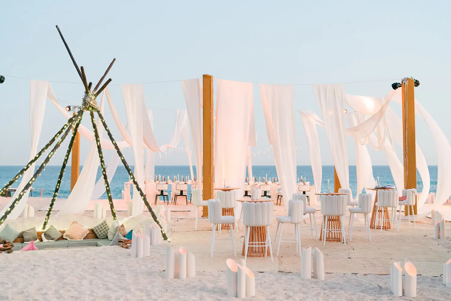 Elegant beach event setup with white drapery, high-back chairs, candlelight, lounge area with pillows, and views of the ocean.