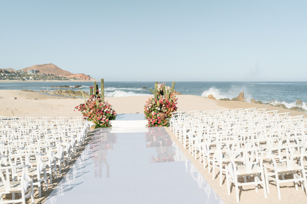 Beach wedding ceremony setup with white chairs, floral arrangements, and cacti lining an aisle to the ocean.