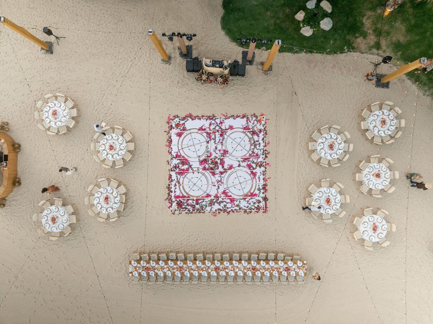 http://Aerial%20view%20of%20a%20beach%20wedding%20reception%20with%20round%20tables,%20a%20floral%20dance%20floor,%20and%20a%20long%20banquet%20table%20in%20the%20sand.