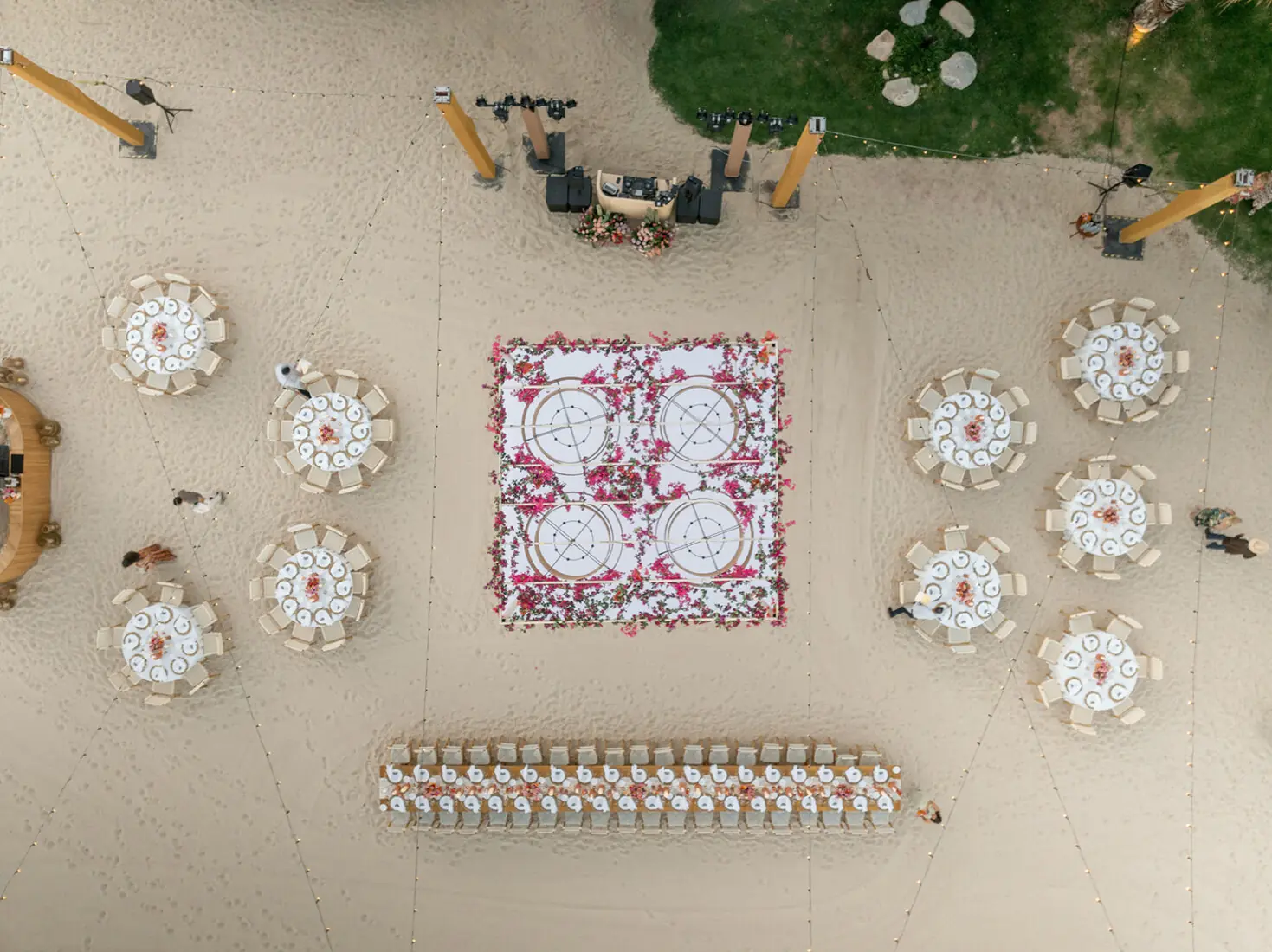 Aerial view of a beach wedding reception with round tables, a floral dance floor, and a long banquet table in the sand.