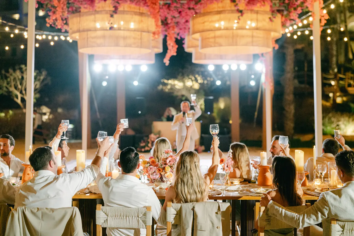 Nighttime wedding reception with guests raising glasses for toast, warm string lights, floral décor, and celebratory atmosphere.