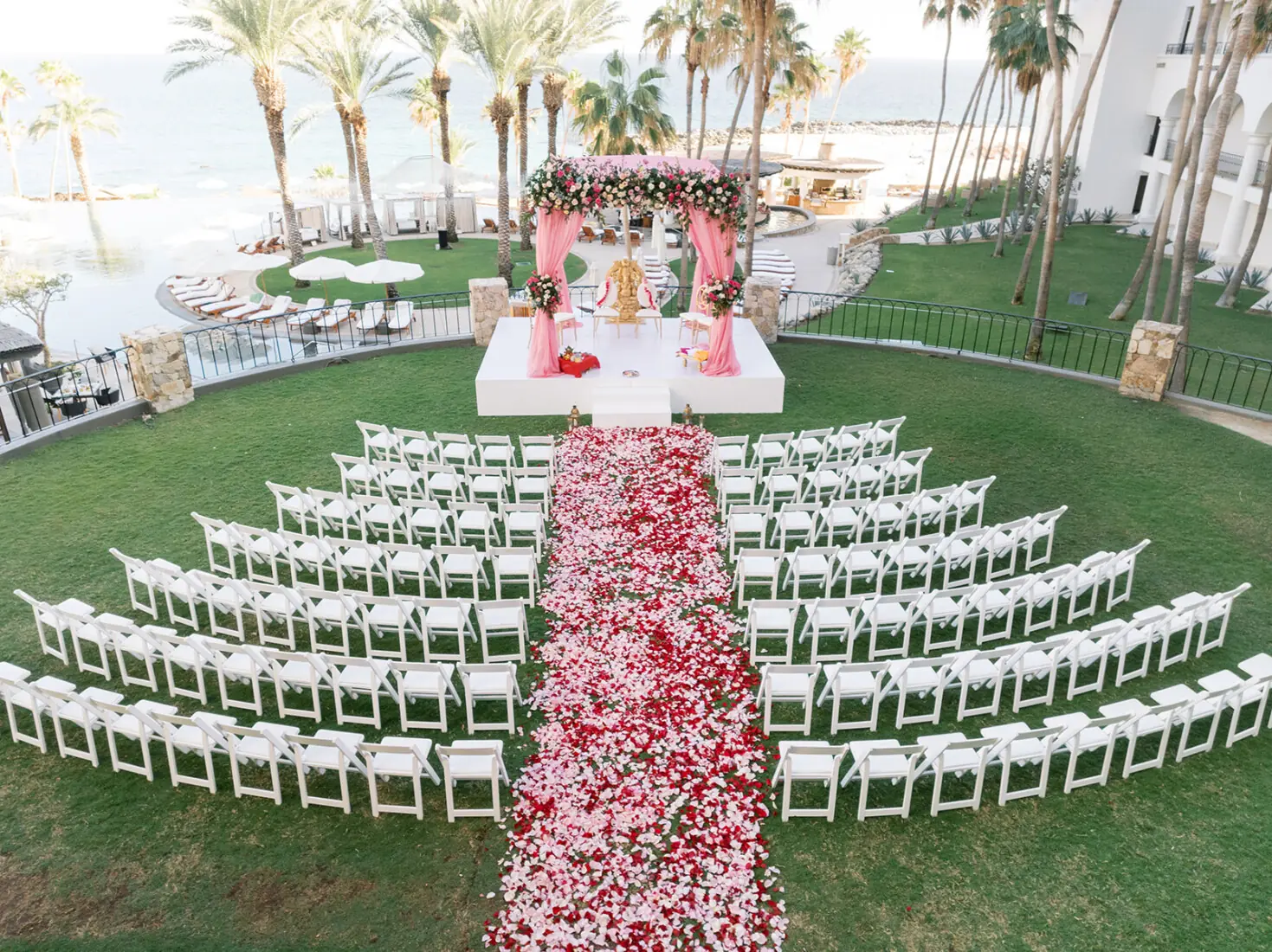 Outdoor wedding ceremony setup with white chairs arranged in semicircles, a stage with floral mandap, and a rose petal aisle, overlooking the ocean.