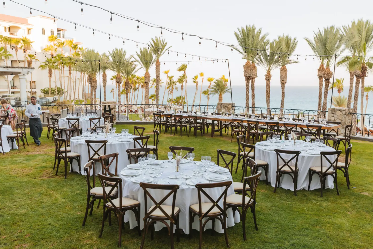 Outdoor wedding reception setup with round tables, string lights, palm trees, and oceanfront backdrop.