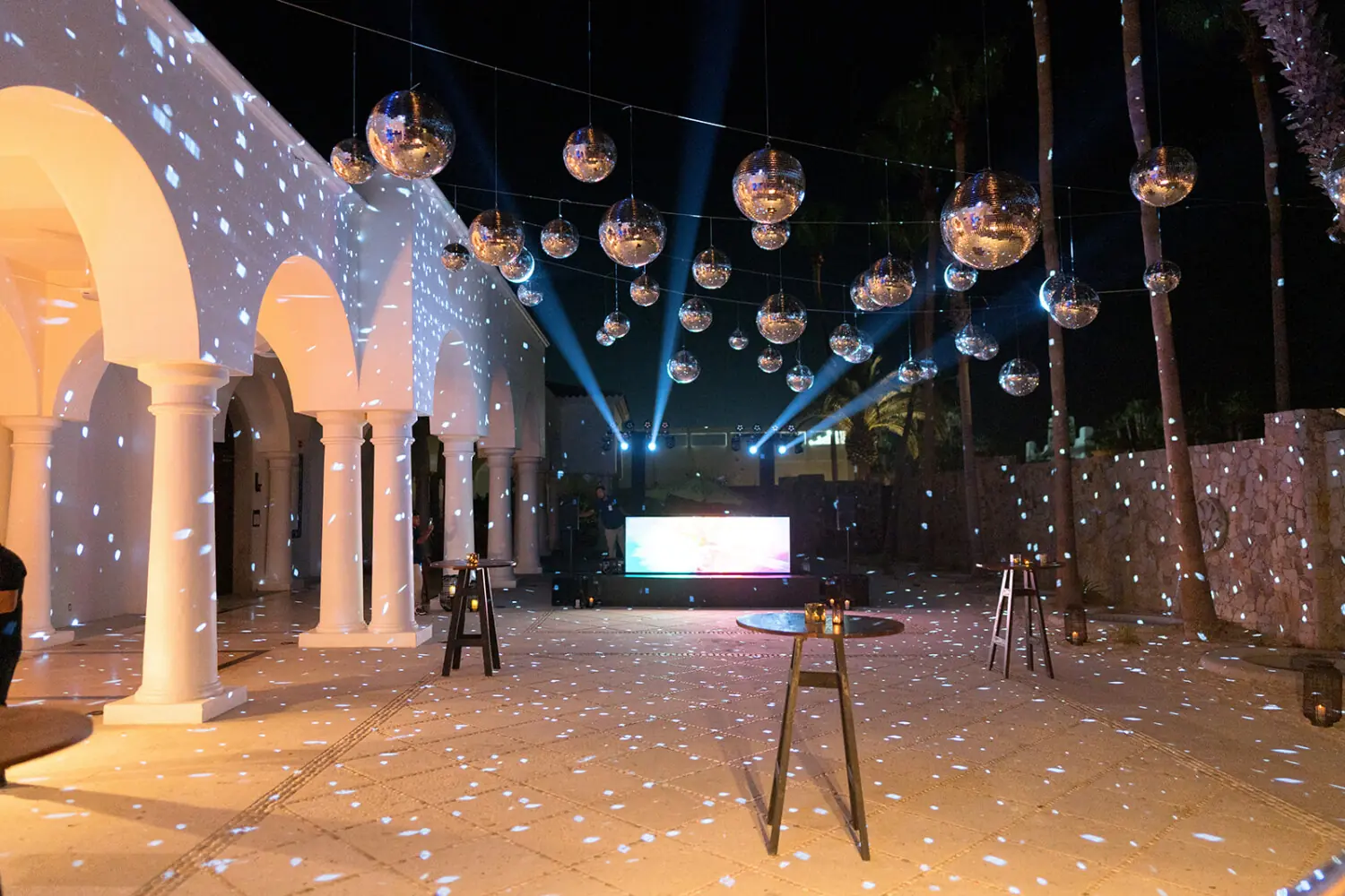 Nighttime event with disco balls overhead, spotlights, and white columns reflecting light on dance floor and walls.