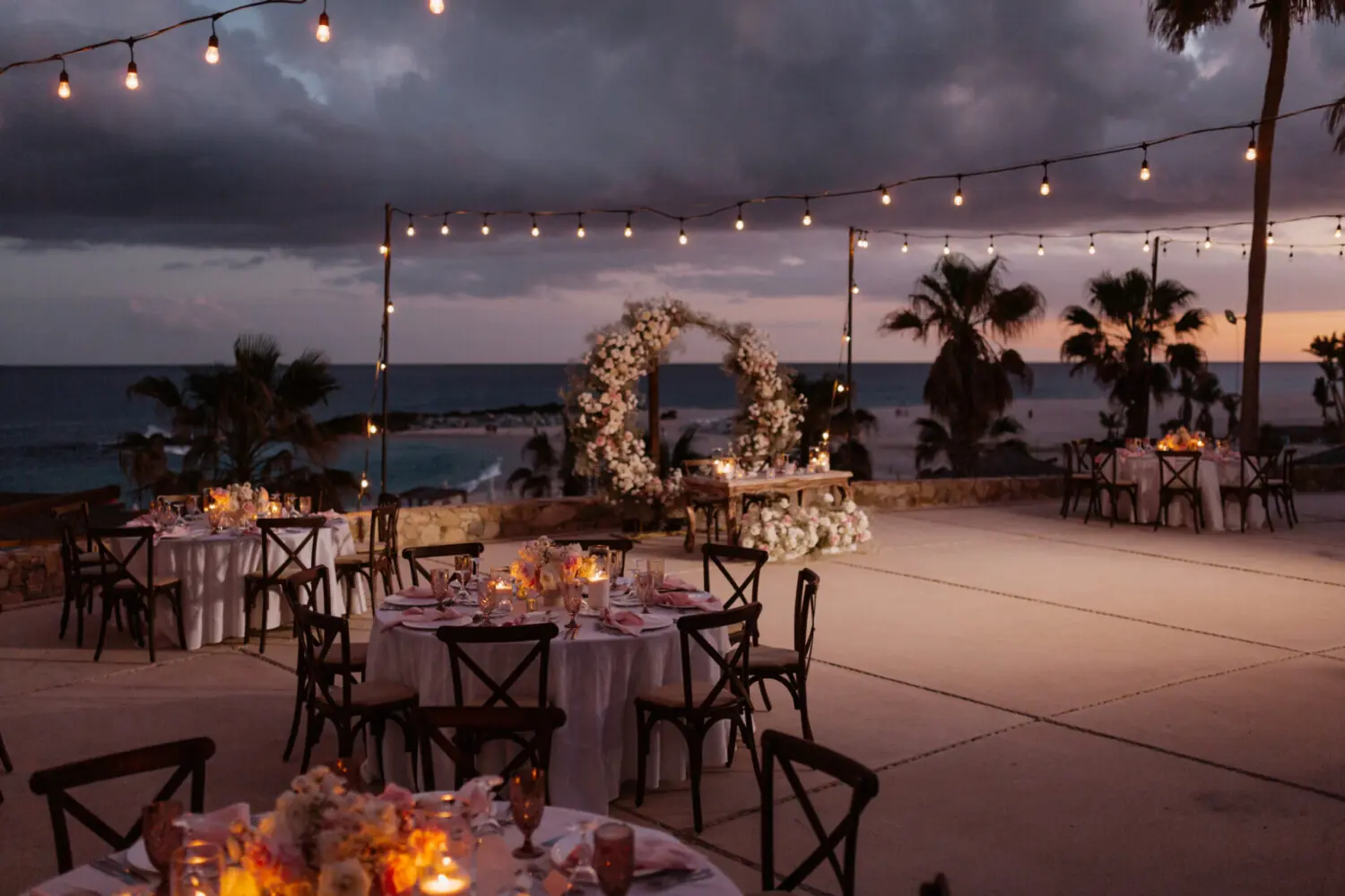 Romantic evening wedding reception with round tables, string lights, floral arch, and ocean view at sunset.