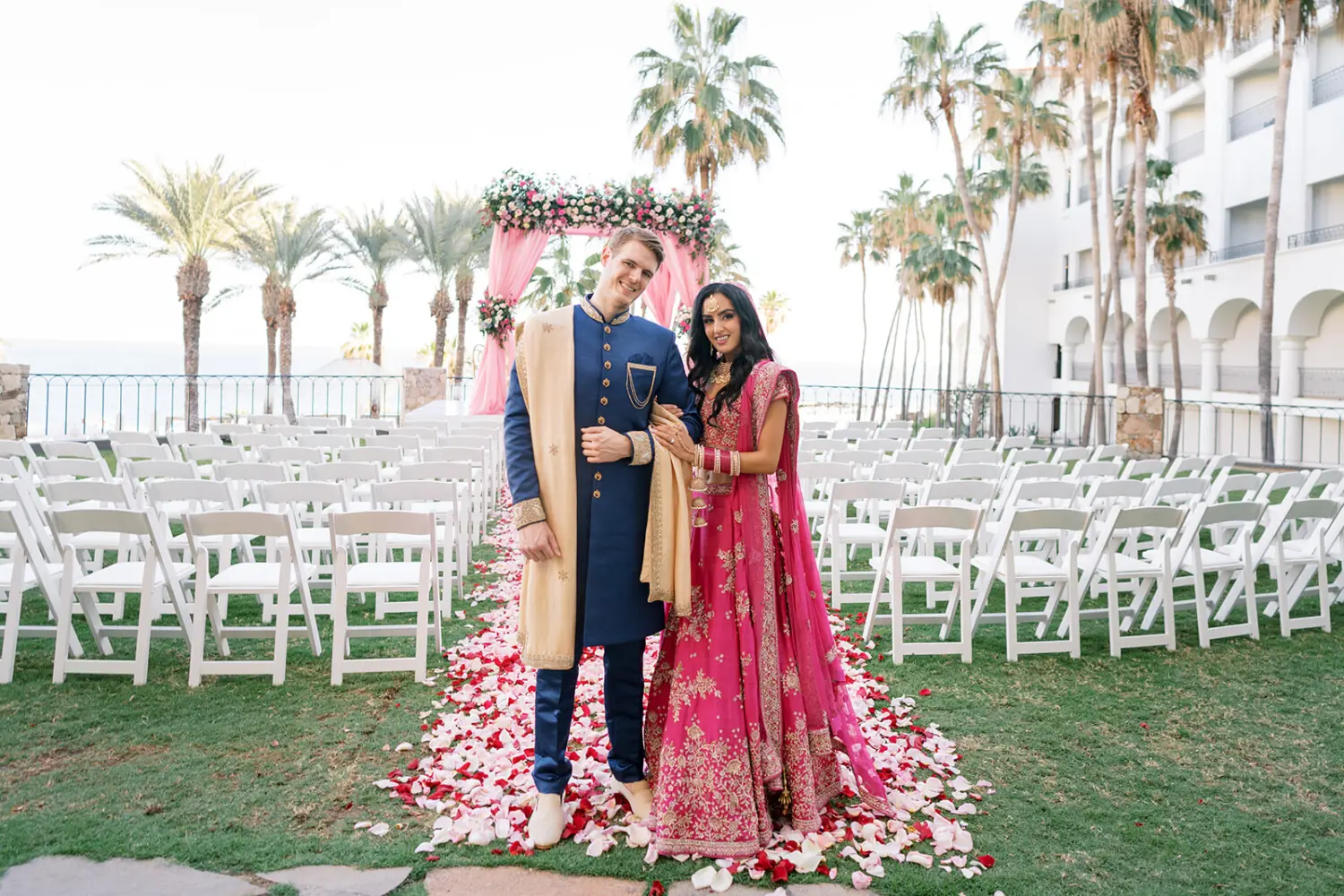 Bride and groom in traditional attire stand on rose petal aisle before floral wedding mandap.