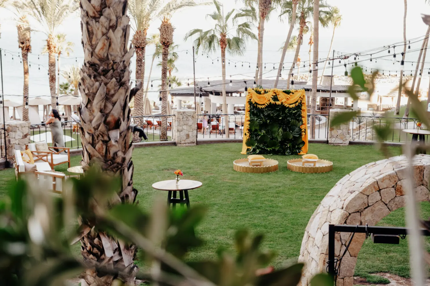 Outdoor event lawn with palm trees, string lights, lounge seating, and decorative yellow floral backdrop.