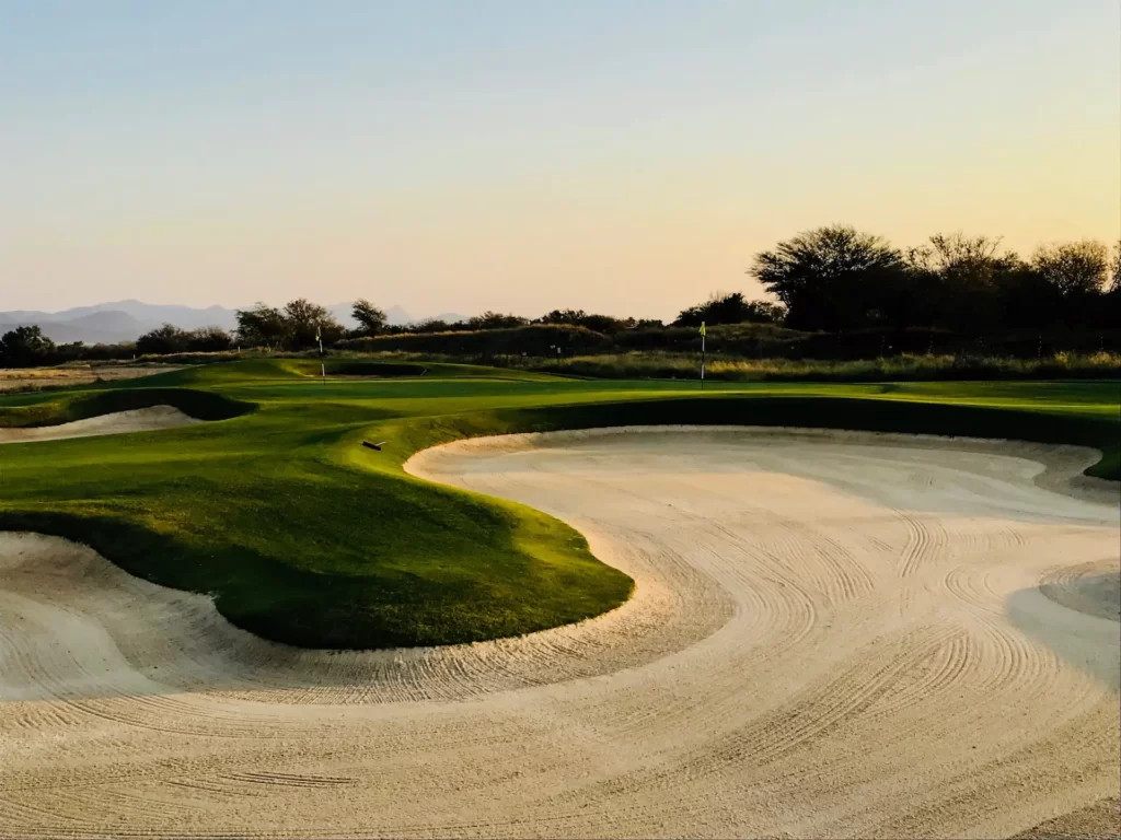 Golf course landscape with raked sand bunkers, green fairways, and distant mountains under warm sunlight.