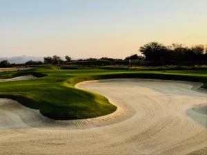 Paisaje de campo de golf con bunkers de arena rastrillados, calles verdes y montañas distantes bajo la cálida luz del sol.