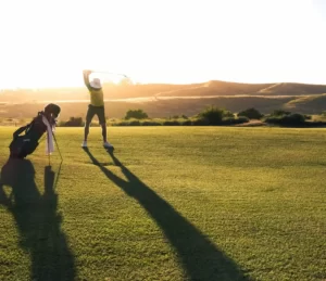 Golfista preparándose para hacer un swing al amanecer en un campo pintoresco con largas sombras y colinas onduladas en el fondo.