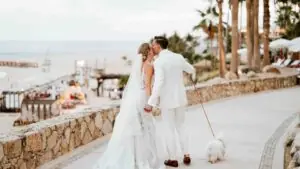 Newlywed couple in white attire kissing on a seaside walkway at sunset, holding hands while walking their small dog.