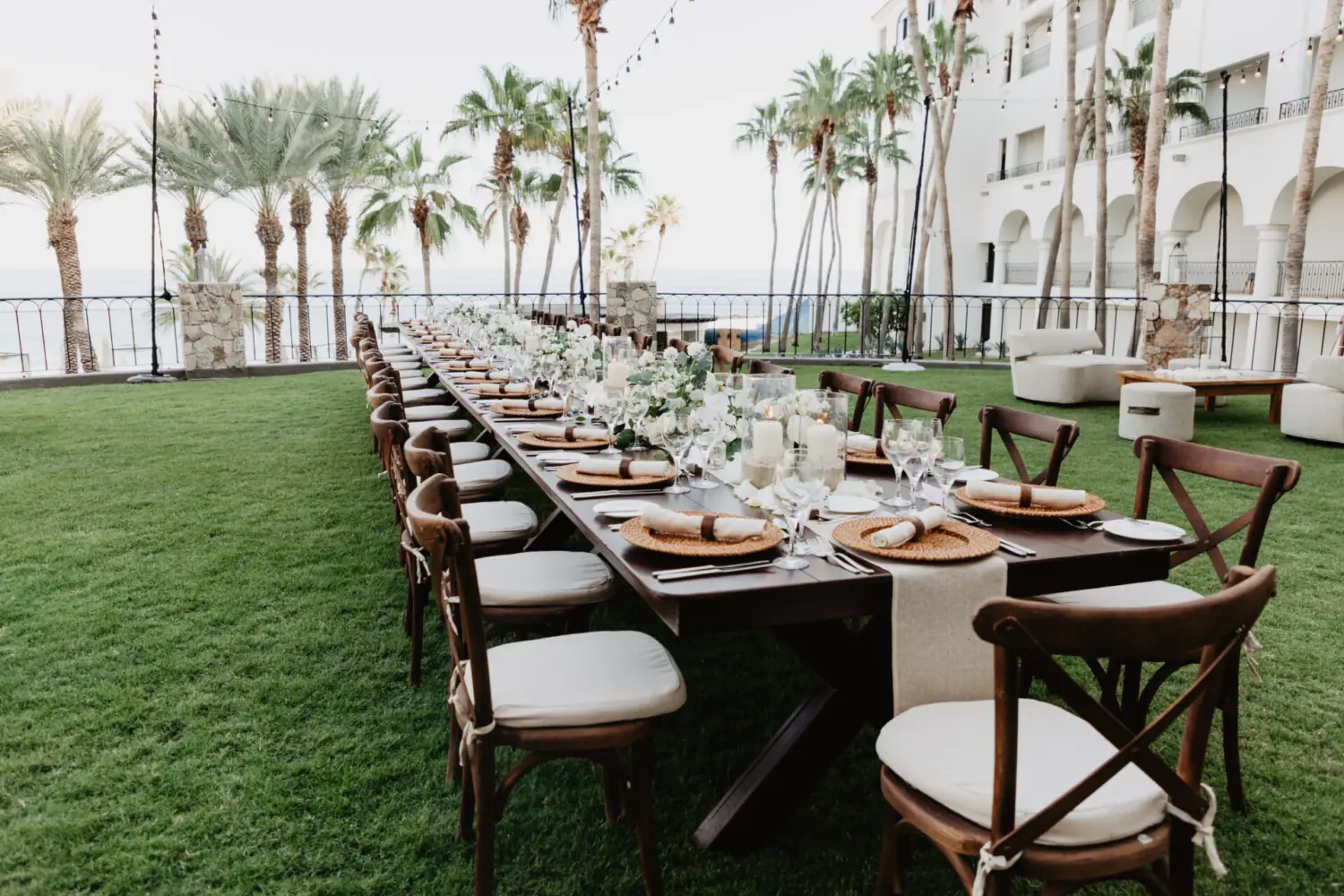 Long outdoor wedding dinner table set with white flowers, candles, woven placemats, palm trees, and ocean view.