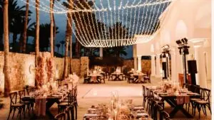 Elegant outdoor wedding reception setup with string lights, decorated tables, palm trees, and romantic evening ambiance.