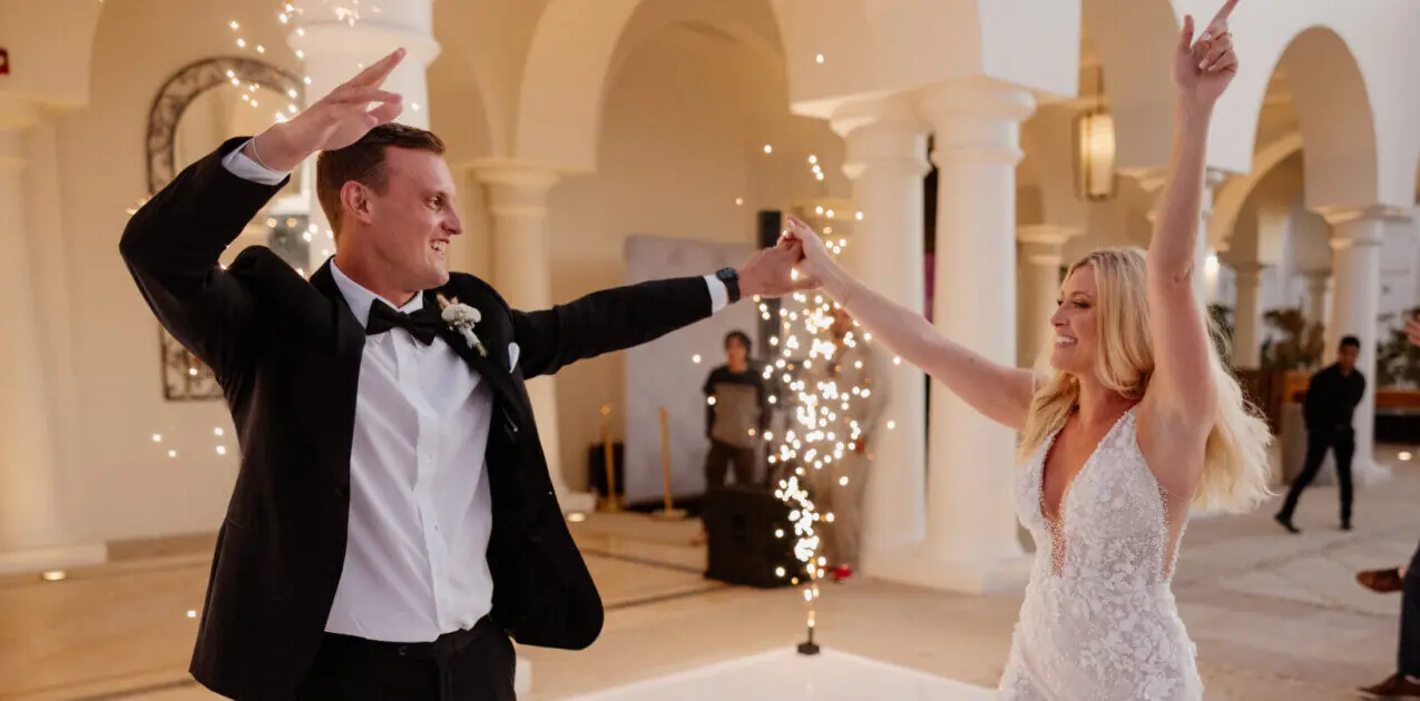 Bride and groom joyfully dance indoors, surrounded by sparklers, smiling while celebrating their wedding together.