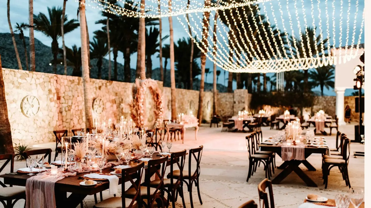 Outdoor wedding reception with string lights overhead, stone walls, decorated tables, candles, and floral arrangements among palm trees.