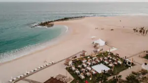 Aerial view of beachside wedding reception with round tables, dance floor, string lights, ocean, and white sand.