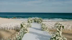White wedding ceremony setup on sandy beach, floral aisle decorations, white chairs, facing turquoise ocean horizon.