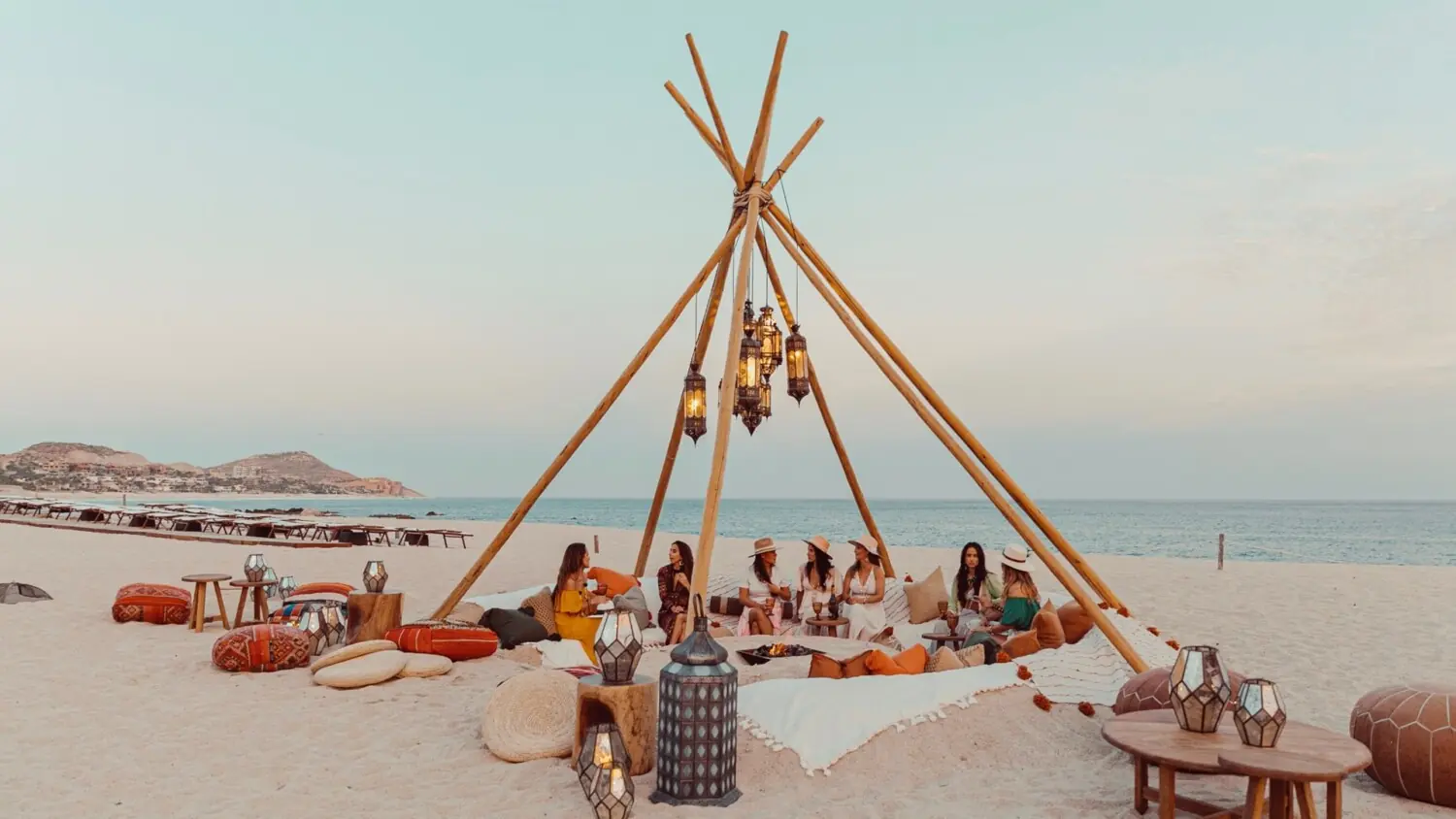 Group of women sit in a boho-style beach setup under a wooden teepee frame with hanging lanterns, surrounded by pillows and ocean views.