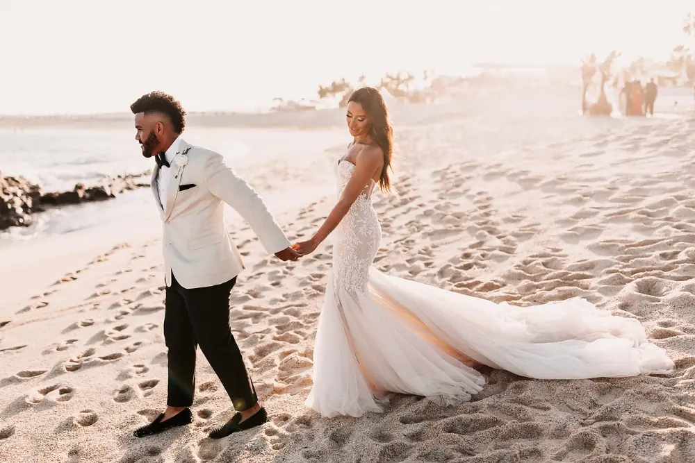 Bride and groom holding hands, walking together across sandy beach, wedding attire flowing, soft sunlight background.
