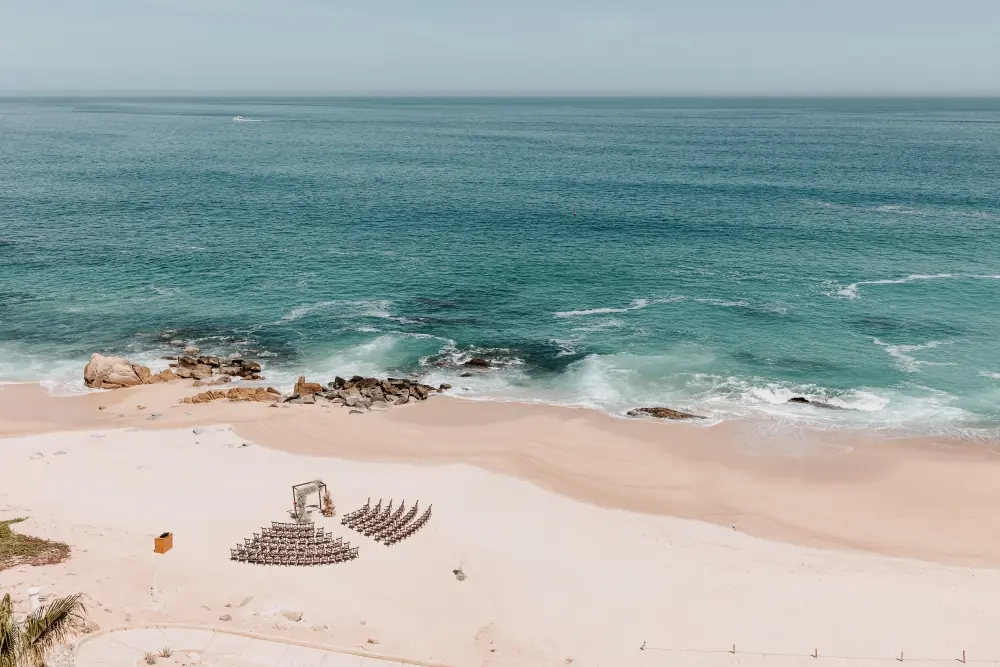 Beach wedding ceremony setup with chairs and arch on sandy shore, turquoise waves and rocks in background.