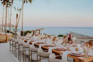 Elegant outdoor wedding reception table with plush chairs, ocean view, candles, glassware, sunset, and palm trees.