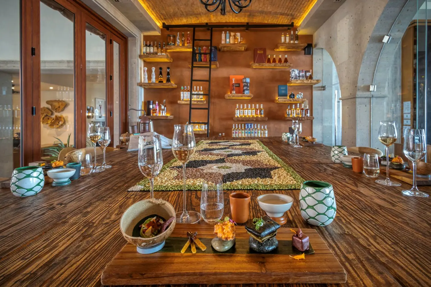 Elegante sala de degustación con mesa de madera, cristalería, cerámica, licores, escalera y estantes decorativos.