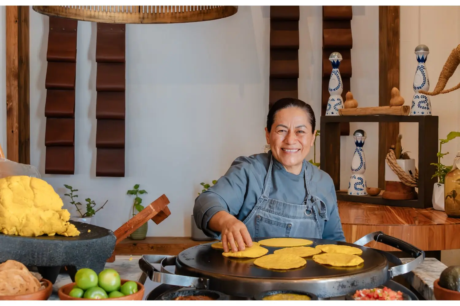Smiling chef prepares fresh corn tortillas on griddle, surrounded by masa, vegetables, and traditional decor.