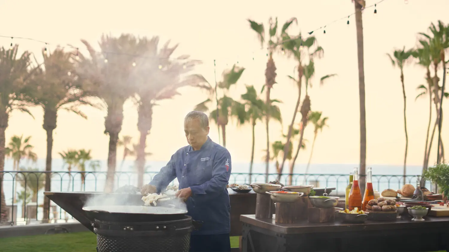 Chef cooks outdoors on large grill with smoke rising, palm trees and ocean in background.