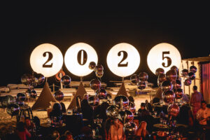 Fiesta nocturna en la playa con carteles luminosos de 2025, globos decorativos y una multitud animada celebrando al aire libre.