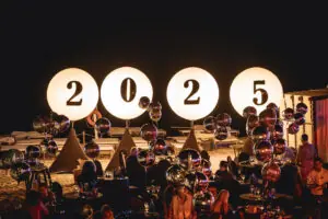 Nighttime beach party with glowing 2025 signs, decorative balloons, and a lively crowd celebrating outdoors.