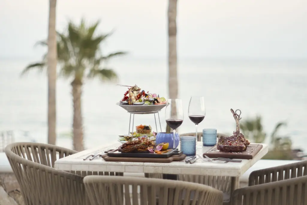 Oceanfront dining table with plated steaks, seafood tower, wine glasses, and palm trees overlooking the sea.