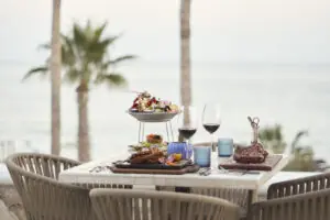 Oceanfront dining table with plated steaks, seafood tower, wine glasses, and palm trees overlooking the sea.