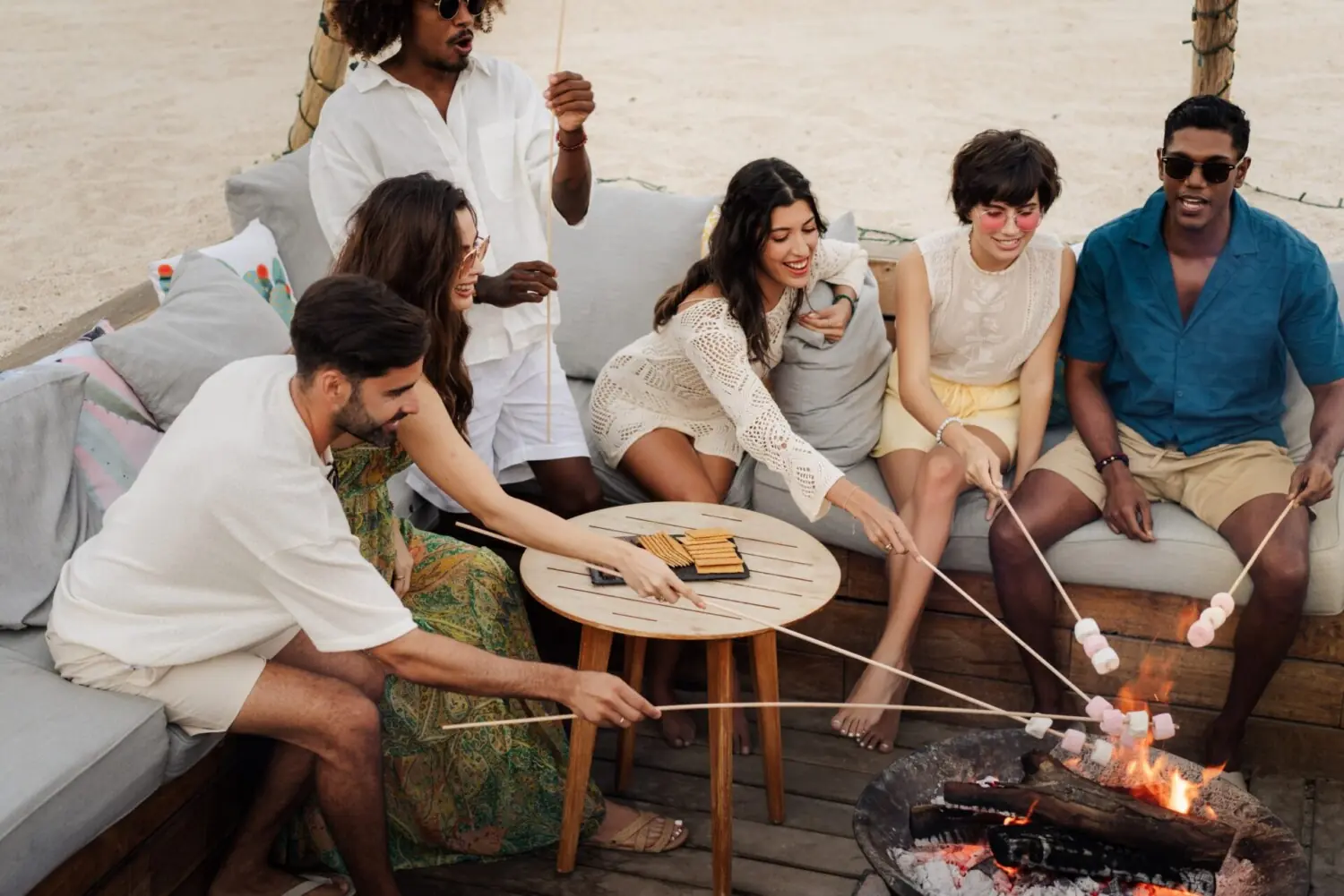 Grupo de amigos sentados juntos al aire libre, asando malvaviscos en una fogata y charlando en un ambiente relajado por la noche.