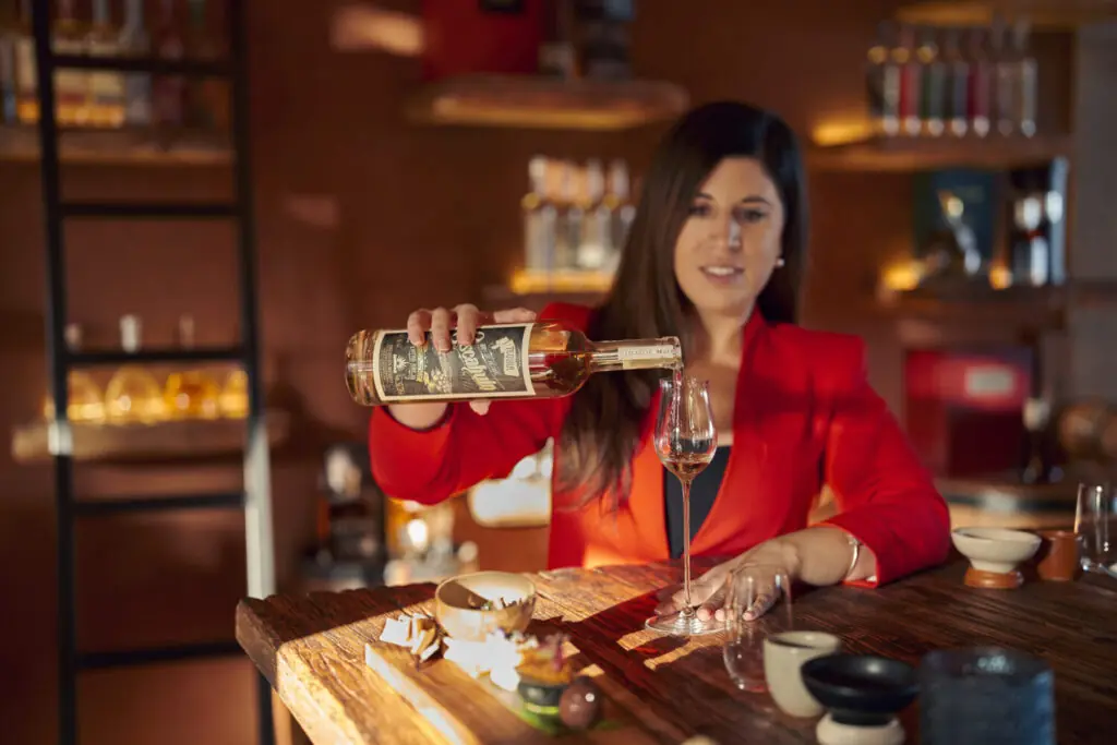Person in a red jacket pouring whiskey into a tasting glass at a wooden bar with bottles displayed behind.