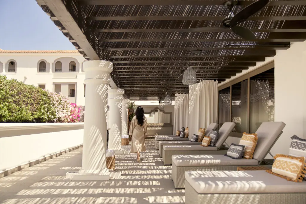 Guest walking through a shaded resort corridor with lounge chairs, white columns, and patterned sunlight.