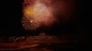Bright fireworks explode in the night sky above ocean shoreline, illuminating water and coastal landscape.