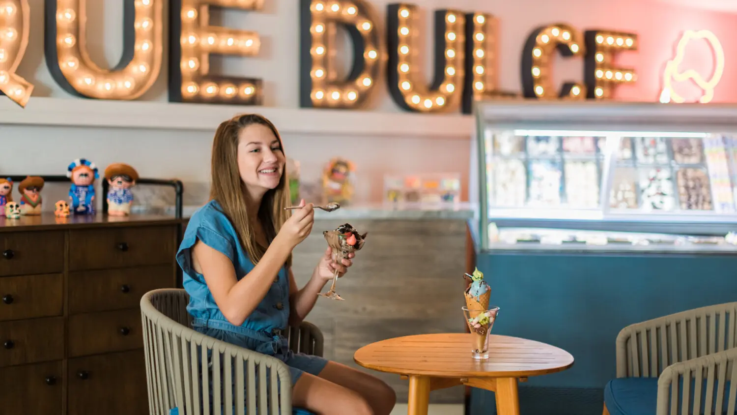 Niña sentada en una mesa en una heladería, comiendo un helado, con una decoración colorida y una exhibición de helados detrás de ella.