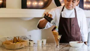 Smiling chef prepares ice cream cone with toppings at counter inside dessert shop.