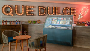 Heladería colorida con letrero de neón "Que Dulce", exhibición de congelador y asientos acogedores.