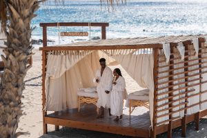 Una pareja con batas blancas se encuentra en una cabaña al aire libre frente a la playa con dos mesas de masaje en el Hilton Los Cabos Beach & Golf Resort.