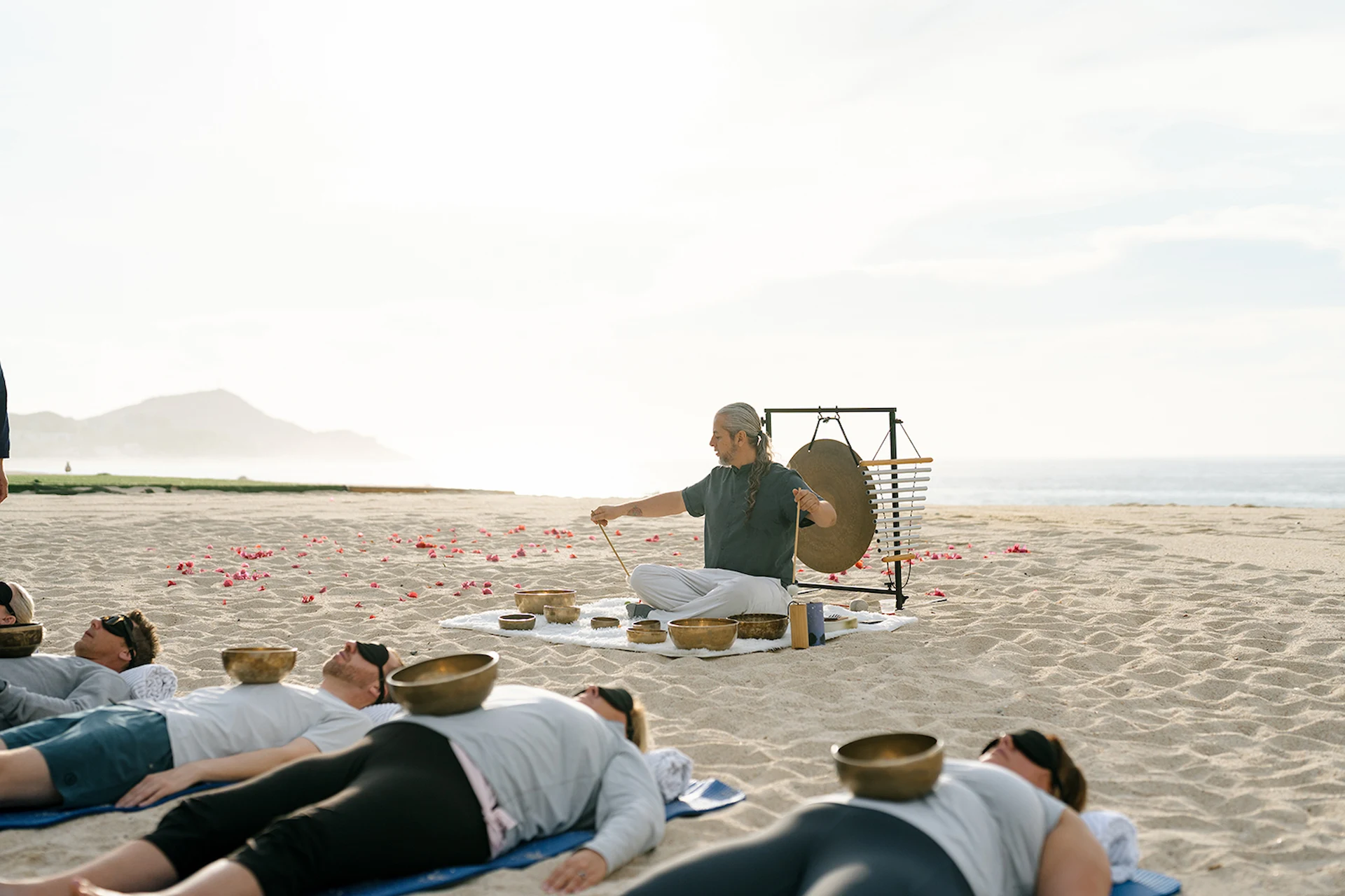 El instructor dirige una sesión de sanación con sonido en una playa de arena, mientras los participantes descansan con cuencos cantores bajo un cielo sereno.