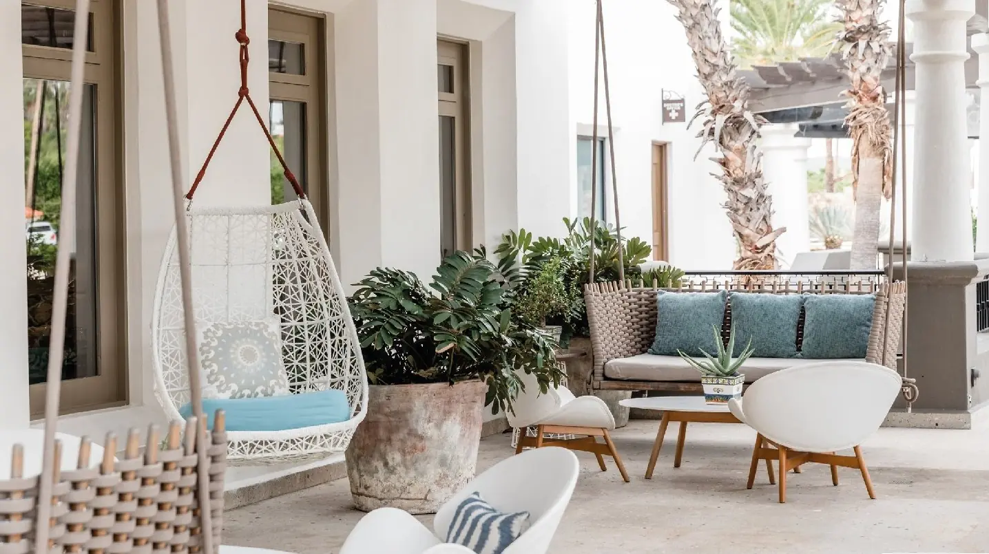 A stylish outdoor lounge with hanging chairs, cushioned seating, potted plants, and palm trees in a covered patio.
