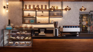 Modern café counter with pastry display case, espresso machine, hanging lights, and stylish shelving decor.