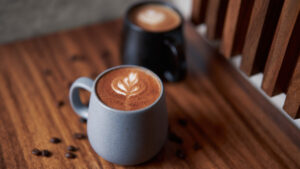 Two mugs of latte art coffee on wooden table, surrounded by scattered roasted coffee beans.